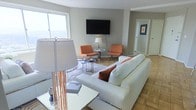 This thoughtfully designed single-floor space offers a clean, contemporary aesthetic that balances comfort and sophistication. The living area serves as the centerpiece, featuring a plush white leather sofa accented with rich burnt-orange throw pillows that echo the warm tones of the matching accent chairs. A wall-mounted flat-screen television anchors one side of the room, while a patterned area rug defines the seating arrangement and adds visual interest to the parquet hardwood flooring beneath. Large windows invite generous natural light, creating a bright and airy atmosphere throughout. A sleek floor lamp with a copper-toned base adds a stylish decorative touch, while a coffee table and side tables provide functional elegance. The neutral gray walls serve as a versatile backdrop, complementing the warm wood tones of the flooring and the bold orange accents of the furniture. A corridor visible from the living area hints at additional rooms beyond, contributing to an overall sense of spaciousness. The space is finished with curated decor details, including framed artwork and carefully chosen accessories, lending the interior a polished, move-in-ready feel.
