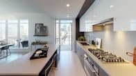 Located in the heart of Downtown Los Angeles, this contemporary residence at 1201 S Hope St offers a sophisticated urban living experience defined by clean lines, premium finishes, and expansive city views. The open-concept layout is anchored by a well-appointed kitchen featuring dark wood cabinetry paired with high-gloss white upper cabinets, a gas cooktop, integrated appliances, and a central island with seating — ideal for both cooking and casual dining. A private balcony is accessible directly from the kitchen, extending the living space outdoors. The living area is flooded with natural light through dramatic floor-to-ceiling windows that frame striking panoramic views of the Los Angeles skyline and surrounding high-rises. Light-toned tile flooring runs throughout, enhancing the bright, airy atmosphere. The bedroom is a serene retreat, finished in soft grey tones with large picture windows that capture sweeping city vistas. Two well-designed bathrooms feature large-format stone tile, deep soaking tubs, walk-in showers, warm wood vanities, and oversized mirrors with recessed lighting. The overall design palette — neutral tones, metallic accents, and modern fixtures — creates a cohesive, refined ambiance throughout this elevated Downtown Los Angeles residence.