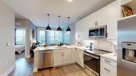 This thoughtfully designed single-floor residence combines modern aesthetics with functional living. The open-concept kitchen is a standout, outfitted with crisp white shaker cabinetry, a bold geometric-patterned backsplash, and a full suite of stainless steel appliances including a French door refrigerator, slide-in range, over-the-range microwave, and dishwasher. A peninsula with a white quartz countertop provides additional workspace and casual seating, while three striking black pendant lights overhead add a contemporary flair. Warm hardwood-style flooring runs throughout the main living areas, seamlessly connecting the kitchen to the living space. Large windows and a glass-paned door flood the living area with natural light and frame sweeping city skyline views, creating an inviting retreat. The bedroom is a serene haven, featuring plush carpeting, a ceiling fan, soft gray accent walls, and generous windows that continue the city views. The bathroom is finished with a wide dual-sink white quartz vanity, matte black hardware, a large frameless mirror, and a sleek walk-in shower. A stacked washer and dryer unit is conveniently tucked nearby, adding everyday practicality to this well-appointed residence.
