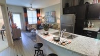 This single-story space showcases an open-concept layout that integrates the kitchen and living areas into one cohesive environment. The kitchen is equipped with stainless steel appliances, including a full-sized refrigerator, and features a curved granite-topped peninsula with a double basin sink and bar seating, making it ideal for casual dining and entertaining. Dark cabinetry above the refrigerator provides additional storage, while the light hardwood flooring extends throughout, unifying the space visually.

The living area is visible directly from the kitchen, creating an inviting, connected atmosphere. A ceiling fan with integrated lighting anchors the seating zone, which benefits from natural light streaming through a large window dressed with bold striped curtains in rust, grey, and black tones. The warm, neutral wall colors are complemented by decorative artwork and ambient table lamps, contributing to a comfortable and stylish ambiance. The thoughtful combination of functional design and modern finishes makes this a practical yet visually appealing single-floor living space.