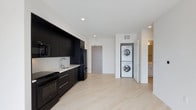 This contemporary single-floor residence showcases a clean, modern aesthetic defined by a striking contrast of dark and light elements throughout. The open-concept kitchen is outfitted with matte black cabinetry that runs floor to ceiling, providing ample storage while delivering a bold, sophisticated look. A suite of black appliances — including a refrigerator, range, and microwave — complements the dark cabinetry, while a white countertop with an undermount sink adds a crisp contrast. Light hardwood-style flooring extends across the main living area, lending warmth and continuity to the space. A dedicated in-unit laundry nook houses a stacked washer and dryer, offering everyday convenience without sacrificing floor space. Recessed lighting in the white ceiling keeps the interior bright and airy. A secondary hallway visible in the background suggests additional living or bathroom space beyond the main area. The overall design is minimal and functional, with clean lines and a neutral palette that creates a polished, move-in-ready environment well-suited for modern living.