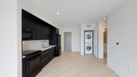 This contemporary single-floor residence showcases a clean, modern aesthetic defined by a striking contrast of dark and light elements throughout. The open-concept kitchen is outfitted with matte black cabinetry that runs floor to ceiling, providing ample storage while delivering a bold, sophisticated look. A suite of black appliances — including a refrigerator, range, and microwave — complements the dark cabinetry, while a white countertop with an undermount sink adds a crisp contrast. Light hardwood-style flooring extends across the main living area, lending warmth and continuity to the space. A dedicated in-unit laundry nook houses a stacked washer and dryer, offering everyday convenience without sacrificing floor space. Recessed lighting in the white ceiling keeps the interior bright and airy. A secondary hallway visible in the background suggests additional living or bathroom space beyond the main area. The overall design is minimal and functional, with clean lines and a neutral palette that creates a polished, move-in-ready environment well-suited for modern living.