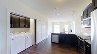 This well-appointed single-floor residence combines modern design with practical convenience in a clean, contemporary layout. The interior showcases rich dark hardwood flooring that runs throughout, creating a warm contrast against the bright white walls and ceilings. The kitchen is a standout feature, outfitted with dark cabinetry, granite countertops, a stainless steel dishwasher, and a central island with an undermount sink. Stylish pendant lights hang above the island, providing focused task lighting while adding a refined aesthetic touch. Large windows at the far end of the kitchen and living area flood the space with natural light, enhancing the open and airy feel. A dedicated in-unit laundry area is thoughtfully integrated into the layout, complete with a full-size washer and dryer tucked behind a set of panel doors with overhead cabinetry for additional storage. Beyond the interior, the space connects to a generous outdoor terrace featuring lush artificial turf, white structural columns, and open railings that frame unobstructed views of the surrounding landscape and waterway. The terrace is well-suited for relaxation or outdoor entertaining under open skies.