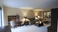 This single-floor space presents a warm and inviting living area defined by its neutral beige walls and soft, light-colored carpeting. The room is illuminated by a combination of wall-mounted uplight sconces and traditional table lamps, which cast a warm, ambient glow across the ceiling and walls, creating a relaxed and intimate atmosphere. Crown molding along the ceiling adds a subtle architectural detail that enhances the overall finish of the space.

The main seating area features a plush sofa and coordinating armchair arranged around a dark wood coffee table, providing a comfortable and well-organized gathering space. Decorative accents, including a vase of fresh flowers and accent pillows, contribute to the room's lived-in charm. A matching end table with an elegant lamp sits beside the sofa, adding both function and style.

Along one wall, a wooden bookcase and a compact secretary desk with a rolling chair define a dedicated workspace or study nook. A decorative globe atop the bookcase adds a classic touch. A framed mirror on the adjacent wall reflects light and contributes to a sense of depth within the space.