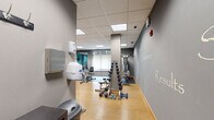 Located at 633 Main St NW in Elk River, Minnesota, this fitness center offers a well-organized and fully equipped exercise space designed to support a comprehensive workout routine. The room features a drop ceiling with recessed panel lighting that provides consistent, bright illumination throughout.

The space is divided into distinct functional zones. The cardio area is outfitted with treadmills, an elliptical trainer, a rowing machine, and stationary bikes, each positioned in front of wall-mounted flat-screen televisions for entertainment during workouts. A large mirror along one wall visually expands the space and aids in form correction. The strength training zone includes a multi-station cable machine, an adjustable weight bench, and a well-stocked dumbbell rack with a range of free weights. Additional accessories such as exercise balls, resistance bands, and step platforms are neatly stored on dedicated racks.

The flooring transitions between warm hardwood-style surfaces in lighter activity zones and durable rubber matting beneath heavy equipment. The walls are painted in bold teal and steel-blue tones, accented by motivational words — including "Strength," "Endurance," "Achieve," and "Results" — applied directly to the walls, reinforcing an energizing atmosphere. A window with a roller shade allows for natural light while maintaining privacy. The overall design is functional, purposeful, and conducive to focused fitness training.