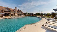 Located at 4285 Julep Way SW in Aiken, South Carolina, this outdoor amenity area is designed to deliver a resort-caliber experience for its residents. The centerpiece is a generously sized, curvilinear swimming pool filled with crystal-clear blue water, surrounded by a broad concrete pool deck that accommodates an extensive array of lounge chairs and shade umbrellas for sun-soaking and relaxation.

A covered cabana structure anchors one end of the pool area, featuring a distinctive metal roof and open-air design that provides a shaded retreat with seating for gatherings. The clubhouse building adjacent to the pool showcases attractive architectural details, including brick accents, a steep gabled roofline with a standing-seam metal roof, and large windows that blend a craftsman-inspired aesthetic with modern sensibility.

The surrounding landscape is carefully curated with ornamental grasses, palm trees, and tropical plantings that reinforce the resort atmosphere. A wrought-iron perimeter fence encloses the space, providing security while maintaining an open, airy feel. Views of the broader residential community are visible beyond the fencing, offering a sense of community integration. The entire outdoor space benefits from abundant South Carolina sunshine, creating a bright, inviting environment ideal for outdoor leisure year-round.