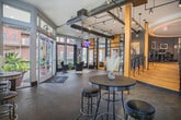 This Memphis lobby presents a striking blend of industrial and contemporary design elements across a single, open floor. The space makes an immediate impression upon entry through glass-paned French doors, which flood the interior with natural light and offer views of the surrounding streetscape and greenery outside.

The flooring throughout features richly toned tile laid in a herringbone pattern, anchoring the space with a warm yet sophisticated character. A defining architectural feature is the sweeping curved staircase at the rear of the lobby, constructed with warm wood treads and elegant wrought iron railings. The staircase frames a circular recessed ceiling detail with ambient lighting, serving as a dramatic focal point.

Exposed black structural beams and ductwork run across the ceiling, complemented by industrial-style track lighting and Edison-bulb pendants that illuminate the space evenly. Steel-framed glass partitions with natural wood accents divide portions of the lobby, adding visual depth and a refined aesthetic.

A comfortable lounge area features plush velvet seating arranged around a gold-accented coffee table beneath large windows dressed with dark curtain panels. A wall-mounted display screen is positioned nearby for added functionality. A high-top bistro table with bar stools offers an additional casual seating option beside the windows. Contemporary artwork decorates the gray-toned walls throughout, completing the polished, modern atmosphere.
