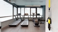 Located at 301 M Street in Washington, DC, this fitness center offers a well-appointed, single-floor exercise facility designed to support a full range of workout routines. The space features rubberized flooring throughout, providing both durability and safety for varied types of training.

The cardio zone is positioned along a wall of large windows that fill the area with natural light, housing treadmills, an elliptical, and stationary cycling bikes. A large display screen is mounted nearby, adding an entertainment option for cardio sessions.

The strength training area is generously equipped, featuring a power rack, cable machines, a full dumbbell rack, medicine balls, stability balls, TRX suspension trainers, and a rowing machine. Multiple strength machines line the perimeter, catering to both upper and lower body training.

Structural columns define distinct workout zones throughout the open layout, while soft blue wall paneling and neutral tones contribute to a calm, focused atmosphere. A mirrored room connects seamlessly from the main floor, offering additional space for stretching or functional training. Recessed and track lighting supplements the natural light, ensuring the space remains bright and energized throughout the day.