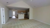 This single-floor home offers an open and airy layout designed for comfortable everyday living. The living area immediately impresses with its soaring vaulted ceiling, which adds a sense of spaciousness and volume to the main gathering space. Soft, neutral wall tones and light carpeting create a warm, welcoming ambiance throughout.

The kitchen is positioned at the heart of the home and is fully equipped with warm-toned wooden cabinetry, stainless steel appliances including a refrigerator and range, and ample counter space. A peninsula with seating provides a natural transition between the kitchen and the living area, making it ideal for casual dining or entertaining. The kitchen features tile or hardwood-style flooring, offering a practical contrast to the carpeted living spaces.

A hallway visible from the main living area leads toward additional rooms and a front entry door, which allows natural light to filter into the interior. The entry area features hard surface flooring, providing a durable transition zone. Recessed lighting throughout the home ensures consistent, functional illumination in every area. Clean white trim and baseboards complement the neutral palette, lending the space a polished, cohesive feel. Overall, the home presents a functional and inviting single-level living environment.