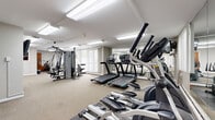 Located at 25151 Brookpark Rd in North Olmsted, Ohio, this dedicated fitness center offers a comprehensive workout environment designed to accommodate a variety of exercise needs. The single-floor space is organized into distinct workout zones, providing ample room for both cardio and strength training activities.

The cardio area is well-equipped with multiple treadmills, elliptical trainers, a stationary air bike, and a recumbent cycle, all positioned to allow for comfortable use. A full-wall mirror spans a significant portion of the room, visually expanding the space and allowing users to monitor their form during exercise. Strength training equipment includes a multi-station cable machine, a leg press, a weight bench, and a dumbbell rack, offering a well-rounded selection for resistance workouts.

The space is finished with neutral-toned carpet flooring and features bright overhead fluorescent lighting throughout. Large windows dressed with vertical blinds allow natural light to filter in and provide views of the surrounding outdoor area. A wall-mounted flat-screen television adds an entertainment element to the workout environment. The overall aesthetic is clean and functional, with white walls and a straightforward layout that maximizes usable floor space for fitness activities.