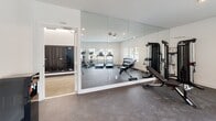 Located at 3810 Gallatin Pike in Nashville, Tennessee, this fitness center is a well-appointed, single-level facility designed to support a full range of workout needs. The main exercise area features durable rubber flooring and is bathed in natural light through multiple large windows fitted with blinds, offering views of the surrounding outdoors. A full wall of floor-to-ceiling mirrors amplifies the sense of space and allows users to monitor their form during workouts. The room is equipped with cardio machines including treadmills and an elliptical, as well as a multi-function cable strength machine, an adjustable weight bench, and a tiered dumbbell rack stocked with a range of free weights. Recessed lighting complements the natural light, keeping the space bright and functional throughout the day. A restroom is included within the facility, finished with large-format stone-look tile extending from floor to wainscot height, providing a clean and polished aesthetic. An adjoining corridor features polished concrete flooring, crisp white walls with dark base trim, and built-in mailbox units, connecting the gym to the broader building with a modern, streamlined design.