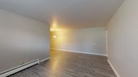 This single-floor unit offers a practical and well-organized layout designed for comfortable everyday living. The open living and dining areas are finished with durable wood-style vinyl flooring in a warm gray tone, complemented by clean white walls and white baseboards that give the space a bright, neutral aesthetic. A pendant light fixture illuminates the dining area, while baseboard heating runs throughout, ensuring warmth across the unit. Natural light filters in through windows fitted with white blinds.

The bedrooms are carpeted in a soft gray tone, providing a cozy contrast to the hard-surface living areas. Each bedroom includes baseboard heating and window-mounted air conditioning units, offering year-round climate control. One bedroom features dual windows, allowing for generous natural light.

The kitchen is efficiently designed with dark cabinetry, light-toned countertops, and a full suite of appliances including a gas range, over-the-range microwave, and a stainless steel sink. A window above the sink adds natural light to the workspace. Open shelving and upper cabinets provide ample storage.

The bathroom is clean and straightforward, featuring a full tub and shower combination with white tile surround, a toilet, and a vanity with sink, all finished with wood-style flooring consistent with the rest of the unit.