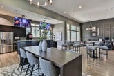 Located in Newnan, Georgia, this single-floor community clubhouse is designed with a contemporary aesthetic that balances comfort and functionality. The open-concept main area features warm hardwood flooring, soaring ceilings, and dramatic dark accent walls adorned with paneling details, creating a sophisticated atmosphere throughout. A statement chandelier anchors the spacious lounge area, which is furnished with plush seating arrangements ideal for relaxing or socializing.

The fully equipped kitchen is a standout feature, outfitted with dark cabinetry, stainless steel appliances, a granite-topped island with bar seating, and a professional-grade coffee station — all complemented by wall-mounted televisions overhead. Bar-height tables and stools are distributed throughout the main gathering area, offering flexible seating configurations for residents.

Private meeting or conference rooms provide a quieter setting, featuring dark walls, large windows that frame verdant outdoor views, wall-mounted displays, and stylish dining-height tables with upholstered chairs — well-suited for focused work or small group gatherings. Throughout the space, large windows invite generous natural light and connect the interior to the surrounding landscape. The overall design blends modern finishes with inviting furnishings, creating a welcoming and versatile community hub.