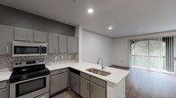 Located along the vibrant South Congress Avenue corridor in Austin's 78704 zip code, this single-story unit presents a clean, contemporary interior designed for functional everyday living. The open-concept layout connects the kitchen and living area seamlessly, creating an expansive, airy feel throughout the main living space.

The kitchen is a standout feature, outfitted with gray flat-front cabinetry, white quartz-style countertops, and a mosaic tile backsplash that adds subtle texture. A peninsula with a stainless steel undermount sink extends into the living area, offering additional prep space and a natural dividing point between the two zones. Stainless steel appliances — including a refrigerator, electric range, over-the-range microwave, and dishwasher — complete the kitchen's modern aesthetic.

Wood-look plank flooring runs throughout the main living areas, providing warmth and durability. The living room receives generous natural light through a large sliding glass door with vertical blinds leading to an outdoor area, complemented by an additional window with horizontal blinds.

The bathroom features a wide vanity with a white basin sink, an expansive wall mirror with a dark trim detail, and a tiled shower-tub combination accented with a classic black-and-white checkered border. Recessed lighting throughout the unit maintains a bright, polished atmosphere.
