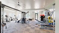Located at 4010 Entrada Dr in Charlottesville, VA, this fitness center offers a generously sized, single-floor workout facility designed to accommodate a full range of exercise needs. The space features a clean, modern aesthetic with crisp white walls, recessed lighting, and a distinctive checkerboard-patterned tile floor that adds visual interest throughout the main training area.

Natural light pours in through large windows lining multiple walls, offering views of the surrounding community and creating a bright, energizing atmosphere. A large ceiling fan overhead aids in air circulation, contributing to a comfortable workout environment.

The facility is thoughtfully organized into distinct zones. A dedicated cardio section houses a row of treadmills, elliptical trainers, and stationary bikes, all positioned along the windows to take advantage of natural light. The strength training area features a multi-station cable machine at the center of the room, along with benches and free weights. A well-stocked dumbbell rack and stability balls are neatly arranged near a wall-mounted flat-screen television.

A separate functional training zone, finished with green rubber flooring, provides space for dynamic exercises and includes a plyo box, suspension trainers, and additional weight equipment. Glass-panel doors lead to an adjacent room, further extending the training options available. The overall layout is open and uncluttered, making it easy to move between zones and equipment.