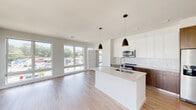 This contemporary single-floor residence is designed with a clean, modern aesthetic throughout. The open-concept living and kitchen area serves as the heart of the space, featuring light wood-tone flooring that extends seamlessly across the entire unit. Large floor-to-ceiling windows line the living area, flooding the interior with abundant natural light while offering views of the surrounding urban streetscape and neighboring buildings.

The kitchen is well-appointed with two-tone cabinetry — crisp white upper cabinets paired with warm walnut-finish lower cabinets — complemented by a white subway tile backsplash and stainless steel appliances including a range and built-in microwave. A central island with a white quartz countertop and undermount sink provides additional workspace and dining space, finished with sleek matte black pendant lights overhead.

A bright, well-proportioned hallway with recessed lighting connects the private areas of the residence. Bedrooms benefit from generous natural light through large windows. The primary bathroom features a glass-enclosed walk-in shower with white tile surround and chrome fixtures, while a second full bathroom offers a tub-shower combination with matching white tile and a walnut-finish vanity. A walk-in closet with wrap-around wire shelving provides ample storage.