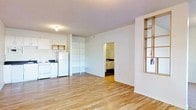 This single-floor space offers a clean, functional layout designed to maximize utility. The main living area features warm-toned hardwood flooring that runs throughout, creating a cohesive and inviting atmosphere. White walls keep the interior feeling bright and open, complemented by flush ceiling lighting.

The kitchen is integrated into the main living space and comes equipped with essential appliances, including a full-size refrigerator and a stovetop range. A small wall-mounted cabinet provides additional storage, while the countertop workspace offers practical room for meal preparation.

A standout feature of the main area is a set of open, built-in shelving units that span from floor to near-ceiling height, offering versatile storage and display options. A distinctive interior window with a light wood frame adds an architectural detail to one wall, allowing light to pass between spaces.

Accessible directly from the main room, the bathroom features dark cabinetry and a well-organized vanity area. The transition between spaces is defined by a clean doorway with a modest threshold, maintaining the open feel of the overall layout.

The space is well-suited for those seeking a straightforward, efficiently organized living environment with all essential areas clearly delineated within a single level.