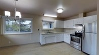 This single-floor space centers around a well-appointed kitchen that combines functionality with a clean, understated aesthetic. The kitchen features white cabinetry that lines both the upper and lower walls, providing ample storage throughout. A classic white tile backsplash extends across the cooking and preparation areas, adding a timeless, easy-to-maintain surface. The stainless steel electric range with oven serves as a focal point along one wall, accompanied by an overhead range hood for ventilation. A double basin sink is positioned beneath a window, allowing natural light to illuminate the workspace while offering an outward view of surrounding greenery and neighboring structures. A pass-through window above the sink connects to an adjacent area, enhancing the open feel of the space. Light hardwood-style flooring runs throughout, contributing warmth and continuity underfoot. Overhead, a flush-mount ceiling light and an under-cabinet fluorescent fixture ensure the kitchen remains well-lit. The overall ambiance is practical and neutral, presenting a move-in-ready environment with a straightforward, functional layout suited to everyday living.