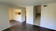 This two-story residence in Midland, Texas offers a straightforward and functional layout across two levels. The main living area on the ground floor is finished with dark hardwood-style flooring that extends throughout, providing a clean and cohesive look. Neutral white walls and a textured popcorn ceiling complement the open feel of the space.

A notable architectural detail on the main level is the built-in pass-through window, which connects the living area to the adjacent kitchen. This practical feature allows for easy communication and flow between the two spaces. The kitchen, partially visible through the pass-through, appears to feature cabinetry and additional built-in shelving.

A carpeted staircase positioned along one wall leads to the second floor, offering a clear transition between levels. The staircase is framed by a clean white wall and includes a white handrail for safety and support.

Overall, the space presents a clean, move-in ready aesthetic with a simple, no-frills design. The combination of dark flooring and white walls creates a neutral palette adaptable to a variety of interior styles.