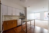 This well-appointed single-floor residence offers a thoughtfully designed open-concept layout that seamlessly connects the kitchen and living areas. The kitchen is a standout feature, showcasing sleek white upper cabinetry paired with warm wood-toned lower cabinets and a textured mosaic tile backsplash. A full suite of stainless steel appliances includes a refrigerator, gas range, over-the-range microwave, and dishwasher, all complemented by light countertops and an undermount sink. Rich hardwood flooring runs throughout the kitchen and entry areas, adding warmth and contrast to the clean, modern aesthetic. A portable island provides additional prep and dining space, enhancing the kitchen's functionality. The open living area transitions to soft, neutral carpeting, creating a comfortable and inviting atmosphere. Large floor-to-ceiling windows flood the space with natural light and frame views of the surrounding urban streetscape, including neighboring residential buildings. The crisp white walls throughout maintain a bright, airy feel. The overall design reflects a contemporary urban sensibility, balancing clean lines, quality finishes, and practical livability in a cohesive and well-lit environment.