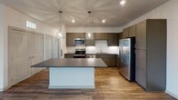 Located on Grandover Village Road in Greensboro, North Carolina, this single-floor residence offers a well-designed layout that balances functionality with modern finishes. The open-concept living and kitchen area is anchored by warm wood-look flooring and recessed lighting, creating a bright, welcoming atmosphere. The kitchen is a standout feature, equipped with taupe shaker-style cabinetry, dark granite countertops, a white subway tile backsplash, and a full suite of stainless steel appliances including a range, microwave, dishwasher, and refrigerator. A central island with pendant lighting provides additional workspace and serves as a natural gathering point. The spacious living area flows seamlessly from the kitchen, offering generous room for various furniture arrangements. Bedrooms feature neutral carpeting and ceiling fans, maintaining a comfortable, understated aesthetic. Large windows fitted with horizontal blinds allow ample natural light while offering views of the surrounding neighborhood and greenery. The bathrooms include a clean, white-tiled walk-in shower with chrome fixtures and grab bars, as well as a separate vanity area with a stone-topped cabinet, undermount sink, and well-lit mirror. Overall, the space presents a clean, contemporary design suited for comfortable everyday living.