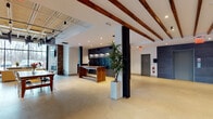 Located at 1409 W Broad St in Quakertown, Pennsylvania, this single-story property showcases a compelling adaptive reuse design that seamlessly merges industrial heritage with contemporary finishes. The spacious lobby serves as the building's centerpiece, featuring exposed wooden ceiling beams, recessed lighting, polished terrazzo flooring, and a bold mailroom wall clad in dark stone tile. A live-edge wood table anchors the entry area, while structural black columns add architectural rhythm throughout.

The lounge area is particularly striking, with floor-to-ceiling windows offering expansive outdoor views, an exposed brick accent wall housing a sleek linear fireplace, a wall-mounted television, and a mix of comfortable modern seating. An adjacent communal dining and gathering space is defined by steel-framed glass partitions, a large farmhouse-style table, and direct sightlines into a stylishly tiled kitchenette.

An on-site fitness center provides a well-equipped workout environment with strength training machines, free weights, cardio equipment, full-length mirrors, and natural light from exterior windows. A dedicated office area features hardwood-style flooring, exposed brick, an open ceiling with visible ductwork, and a large window with outdoor views. An elevator clad in dark stone tile provides accessibility throughout the building.