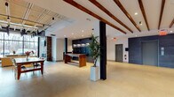Located at 1409 W Broad St in Quakertown, Pennsylvania, this single-story property showcases a compelling adaptive reuse design that seamlessly merges industrial heritage with contemporary finishes. The spacious lobby serves as the building's centerpiece, featuring exposed wooden ceiling beams, recessed lighting, polished terrazzo flooring, and a bold mailroom wall clad in dark stone tile. A live-edge wood table anchors the entry area, while structural black columns add architectural rhythm throughout.

The lounge area is particularly striking, with floor-to-ceiling windows offering expansive outdoor views, an exposed brick accent wall housing a sleek linear fireplace, a wall-mounted television, and a mix of comfortable modern seating. An adjacent communal dining and gathering space is defined by steel-framed glass partitions, a large farmhouse-style table, and direct sightlines into a stylishly tiled kitchenette.

An on-site fitness center provides a well-equipped workout environment with strength training machines, free weights, cardio equipment, full-length mirrors, and natural light from exterior windows. A dedicated office area features hardwood-style flooring, exposed brick, an open ceiling with visible ductwork, and a large window with outdoor views. An elevator clad in dark stone tile provides accessibility throughout the building.