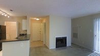 This single-floor residence offers a practical and comfortable layout with a warm, neutral aesthetic throughout. The main living area is carpeted and centers around a fireplace with a white surround and glass-front screen, serving as a natural focal point in the room. A wall-mounted vent nearby suggests a climate-control system integrated into the space. The textured ceiling and light-toned walls contribute to a clean, understated look throughout.

Adjacent to the living area, the entryway features tile flooring and a white front door accompanied by a louvered bifold closet, providing convenient storage upon entry. A ceiling-mounted light fixture illuminates the foyer, creating a welcoming transition into the home.

The kitchen, partially visible from the living space, includes white cabinetry, a dark countertop, and a sink positioned along the perimeter. The open sightlines between the kitchen and living room promote a connected, functional flow. Overall, the space presents a straightforward and livable environment with classic residential finishes and practical amenities suited for day-to-day use.