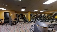 This fitness center offers a comprehensive workout environment designed to meet a variety of exercise needs. The space is outfitted with an array of cardio machines, including treadmills, ellipticals, and upright bikes from brands such as Octane, positioned along one side of the room for easy access. Complementing the cardio equipment is a dedicated strength training area featuring free weights, a weight bench, and cable machines, providing options for both resistance and functional training. The room is brightened by recessed fluorescent panel lighting set into a drop-tile ceiling, ensuring a well-lit atmosphere throughout. Warm yellow-toned walls with accent columns give the space a welcoming feel, while full-length mirrors along one wall enhance the sense of openness and allow users to monitor their form. The floor is covered in a bold, multicolored wave-patterned carpet that adds a lively visual element to the room. The layout is thoughtfully organized, with equipment spaced to allow comfortable movement between stations. Overall, this fitness center delivers a functional and energetic environment suited for a full range of workout routines.