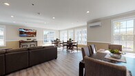 Located at 189 Paddlers Point in Milford, PA, this single-floor community space is thoughtfully designed to accommodate gathering, dining, and everyday socializing. The layout is open and generously proportioned, with distinct functional zones that flow seamlessly into one another.

The main gathering area features a large sectional leather sofa anchoring a comfortable lounge space, complemented by recessed lighting and crown molding that lend a polished, welcoming ambiance. Light wood-tone flooring runs throughout, contributing to a cohesive and clean aesthetic. A wall-mounted mini-split system provides climate control.

Multiple dining areas are arranged throughout the space, each furnished with dark wood tables and chairs suited for group use. Large double-hung windows line the perimeter walls, flooding the interior with natural light and offering views of the surrounding grounds and neighboring buildings. The two-tone wall treatment—neutral upper walls paired with warm tan wainscoting—adds visual structure throughout.

The kitchen is well-appointed with white shaker cabinetry extending to the ceiling, granite countertops, and a full suite of stainless steel appliances including a range, microwave, and dishwasher. A separate room with additional seating and a wall-mounted television provides a more intimate setting within the overall layout.