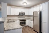 This single-floor residence offers a thoughtfully designed open-concept layout that seamlessly connects the kitchen and living areas. The kitchen is well-equipped with white shaker-style cabinetry, granite-look countertops, stainless steel appliances including a refrigerator, range, microwave, and dishwasher, and a peninsula that provides additional counter space and separates the kitchen from the living area. The living space features durable wood-look flooring and a ceiling fan, creating a comfortable and functional environment. Baseboard heating is present throughout, ensuring efficient climate control. The bedrooms are carpeted for warmth and comfort, each featuring ceiling fans and generous closet space with built-in hanging rods. The bathroom is finished with a granite-topped white vanity, a large mirror, white cabinetry for storage, and a full shower-tub combination. A dedicated entry area with wood-look flooring provides a welcoming transition into the main living space. The attached garage offers a spacious, clean concrete-floored area with an automatic opener, providing secure parking and additional storage. The overall aesthetic is clean and neutral, with light-toned walls throughout that lend a bright, move-in-ready appeal.