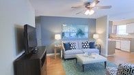 This single-floor residence offers a well-appointed living space designed with a cohesive and inviting aesthetic. The living room serves as the centerpiece of the home, showcasing warm hardwood-style flooring that contrasts beautifully with the cool, calming blue accent wall. A large statement artwork in complementary greens and blues anchors the space, adding a touch of contemporary sophistication. The room is furnished with a light-toned sofa accented by navy and polka-dot throw pillows, paired with a tufted ottoman that doubles as a coffee table. Mirrored side tables flank the seating area, each topped with elegant table lamps that provide warm ambient lighting. A ceiling fan with an integrated light fixture adds both functionality and a casual charm to the room. A dark media console hosts a flat-screen television, grounding one side of the space. A teal patterned area rug ties the furniture arrangement together, adding texture and depth to the floor. The overall ambiance of the space is fresh, modern, and comfortable, with a carefully curated color palette of blues, grays, and neutrals that creates a serene and welcoming atmosphere throughout.