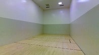 This single-level space is a dedicated racquetball or squash court designed for active recreation. The court features a smooth, light-toned hardwood floor finished with clearly marked regulation playing lines in red and yellow, providing players with precise boundary references during gameplay. The floor's natural wood grain is well-preserved, reflecting the overhead lighting and contributing to the court's clean, functional aesthetic.

The surrounding walls are finished in a two-tone treatment: a lighter white upper section and a slightly greener lower panel, which is a standard design feature that helps players distinguish the playable wall surface. The walls appear smooth and well-maintained, essential for consistent ball bounce and rebound performance.

Overhead, the court is equipped with recessed fluorescent lighting panels that provide even, shadow-minimizing illumination across the entire playing surface. Ventilation is provided through ceiling-mounted HVAC vents, ensuring adequate airflow during intense play sessions. The fully enclosed design with tall ceilings creates an immersive, distraction-free environment well-suited for competitive and recreational play alike.