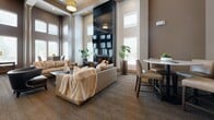 The Aksarben Clubroom in Omaha, Nebraska is a thoughtfully designed community space that blends comfort and sophistication across a single, generously sized floor. The main lounge area commands immediate attention with its soaring double-height ceilings, dramatic floor-to-ceiling tiled fireplace, and a wall-mounted television, creating an inviting focal point for relaxation and socializing. Large, multi-pane windows flood the space with abundant natural light while elegant structural columns add a refined architectural character. Plush neutral-toned sofas and armchairs are arranged throughout multiple seating zones, complemented by glass-topped accent tables and gold-framed open shelving units that lend a contemporary, upscale feel. A dedicated dining area features high-top round tables with leather bar-height chairs, ideal for casual gatherings. The fully equipped kitchen boasts rich dark wood cabinetry, a herringbone tile backsplash, stainless steel appliances including a full-size refrigerator, and a breakfast bar with stylish pendant lighting overhead. Warm taupe walls, plush carpeting, and decorative greenery throughout contribute to a cohesive and welcoming ambiance, making this clubroom a versatile and elegant venue for community events and everyday use.