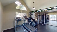 Located at 10951 Laureate Dr in San Antonio, TX, this single-story fitness center offers a well-equipped workout facility designed to support a comprehensive exercise routine. The space is defined by striking vaulted ceilings that rise to a dramatic peak, creating an open and airy atmosphere throughout. A signature arched window near the roofline floods the interior with natural light, complemented by additional rectangular windows that frame views of the surrounding trees and community exterior.

Pendant-style lighting fixtures are suspended from the high ceiling, providing functional illumination across the rubber-floored workout area. The cardio section is outfitted with multiple treadmills, a StairMaster step climber, and an elliptical machine, all arranged to take advantage of the natural light and outdoor views. A dedicated strength training zone features a multi-station cable machine system, along with a rack of dumbbells in varying weights and a workout bench for versatile training options.

A wall-mounted flat-screen television adds an entertainment element to the space, while a decorative neon sign reading "Good Things Come To Those Who Sweat" runs along a floral garland display, adding personality and motivation to the environment. Exercise balls are neatly stored on a rack near the entrance. A set of glass sliding doors and a secondary door provide direct access to the exterior, enhancing both convenience and ventilation throughout the facility.