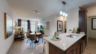 This single-floor residence showcases a thoughtfully designed open-concept layout that seamlessly connects the kitchen, dining, and living areas. The kitchen is anchored by a central island with granite countertops, rich dark-wood cabinetry, and stainless steel appliances, including a French door refrigerator and built-in microwave. Pendant lighting above the island adds a polished touch, while wood-style flooring extends throughout the main living areas.

The dining space accommodates a round table with modern chairs, positioned beneath track lighting and adjacent to large windows dressed in neutral blinds and dark curtains, allowing ample natural light. The living room features neutral-toned walls, a geometric mirror wall arrangement, and generous window coverage that enhances the bright, airy ambiance.

The bedroom offers plush carpeting, large windows for natural light, and direct access to the en-suite bathroom. The bathroom includes a dual-sink vanity with granite countertops, dark wood cabinetry, a large mirror, and tile flooring. A dedicated laundry area houses side-by-side washer and dryer units with overhead wire shelving, adding practical utility to the residence's well-appointed design.