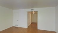 This single-floor space is characterized by its clean, minimalist design and practical layout. The main living area features warm-toned hardwood flooring that extends throughout, providing a cohesive and inviting foundation. Crisp white walls and a smooth white ceiling create a bright, open atmosphere, enhanced by overhead HVAC vents that ensure consistent climate control throughout the space.

A wide opening connects the main living area to a hallway that leads directly to the unit's entry door, which is fitted with a ceiling-mounted flush light fixture that illuminates the corridor. The hallway features the same hardwood flooring as the living area, maintaining visual continuity. Along the hallway, multiple interior doors provide access to additional rooms, offering a well-organized and private layout.

The living area includes a flush baseboard vent along the lower wall, contributing to efficient airflow. The walls are free of ornamental detail, keeping the aesthetic simple and versatile. Overall, the space presents a functional and tidy environment with a neutral palette, making it well-suited for a variety of furnishing styles and personal preferences.