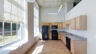 This single-floor space showcases an industrial loft aesthetic, characterized by dramatically high ceilings with exposed painted wood planking and visible ductwork that add architectural character. The kitchen is a standout feature, designed in an L-shaped layout and outfitted with light natural wood cabinetry that provides generous storage across both upper and lower units. Black appliances, including a full-size refrigerator, gas range with overhead microwave, and dishwasher, contrast cleanly against the light cabinetry and grey countertops. A double sink is positioned along the windowed wall, offering natural light while working. The space benefits from an abundance of natural light streaming through tall, arched windows along one wall, casting bright patches across the light hardwood flooring. The open, airy atmosphere is further enhanced by the considerable ceiling height, making the kitchen and living area feel expansive. Exposed utility pipes run along the upper walls, reinforcing the building's industrial character while maintaining a clean, functional interior. The overall ambiance is bright, open, and utilitarian with a modern edge.