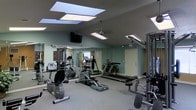 This fitness center is a thoughtfully designed, single-floor workout facility that offers a comprehensive exercise environment. The room features vaulted ceilings with multiple skylights that allow natural light to flood the space, complemented by a ceiling fan that promotes air circulation during workouts. Recessed and panel lighting ensures the area remains well-lit at all times.

The facility is outfitted with a diverse selection of cardio equipment, including treadmills and an elliptical trainer, as well as a multi-station cable strength training machine, providing users with a full range of exercise options. The equipment is arranged to maximize usable floor space while maintaining clear pathways throughout the room.

Full-length mirrors line one wall, visually expanding the space and allowing users to monitor their form during exercise. The walls are painted in a soft, muted green tone that contributes to a calm and focused atmosphere. A wall-mounted flat-screen television adds an entertainment element for those looking to stay engaged during longer workout sessions.

The flooring consists of a neutral, low-pile carpet suited for gym use, and a potted plant near the corner adds a touch of natural warmth to the utilitarian setting. A private entrance door at the rear of the room provides convenient access to the facility.