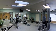 This fitness center is a thoughtfully designed, single-floor workout facility that offers a comprehensive exercise environment. The room features vaulted ceilings with multiple skylights that allow natural light to flood the space, complemented by a ceiling fan that promotes air circulation during workouts. Recessed and panel lighting ensures the area remains well-lit at all times.

The facility is outfitted with a diverse selection of cardio equipment, including treadmills and an elliptical trainer, as well as a multi-station cable strength training machine, providing users with a full range of exercise options. The equipment is arranged to maximize usable floor space while maintaining clear pathways throughout the room.

Full-length mirrors line one wall, visually expanding the space and allowing users to monitor their form during exercise. The walls are painted in a soft, muted green tone that contributes to a calm and focused atmosphere. A wall-mounted flat-screen television adds an entertainment element for those looking to stay engaged during longer workout sessions.

The flooring consists of a neutral, low-pile carpet suited for gym use, and a potted plant near the corner adds a touch of natural warmth to the utilitarian setting. A private entrance door at the rear of the room provides convenient access to the facility.