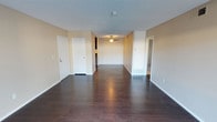This single-floor space presents a clean, open layout highlighted by rich, dark hardwood flooring that extends throughout the main living area, creating a sense of continuity and warmth. The neutral beige walls provide a versatile backdrop suitable for a variety of décor styles. A ceiling fan with a built-in light fixture is centrally positioned, offering both air circulation and ambient lighting to the space.

The living area is generously proportioned, with a long, open floor plan that allows for flexible furniture arrangement. White-painted doors and trim provide crisp contrast against the warm wall tones. A floor vent indicates a forced-air climate control system integrated into the space.

Toward the rear of the main area, a glimpse into the kitchen reveals dark cabinetry, hinting at additional functional space beyond the living room. A doorway to the right leads to additional rooms, suggesting a practical and connected layout throughout the unit. The textured ceiling adds a traditional finish to the overall interior. The space is vacant, allowing prospective occupants to clearly assess its layout and potential.