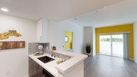This contemporary single-floor space makes a strong visual impression through its clean, modern design and thoughtful use of color. The kitchen is a standout feature, showcasing crisp white cabinetry, dark lower cabinets for contrast, and a smooth white countertop that extends into a peninsula with an undermount sink. A mosaic tile backsplash in neutral grey tones adds texture and sophistication to the cooking area, complementing the overall modern aesthetic.

The open layout allows for a seamless flow between the kitchen and the adjoining living areas. Wide-plank hardwood-style flooring runs throughout, grounding the space with warmth and continuity. White walls and ceilings maximize the sense of brightness and openness, while bold yellow accent walls punctuate the interior with striking pops of color, visible through doorways that connect the various areas of the space.

Natural light plays a key role in the ambiance, with a glazed entry door inviting sunlight deep into the interior and offering a glimpse of lush greenery beyond. Decorative touches such as potted plants add a welcoming, lived-in quality. The overall atmosphere is fresh, vibrant, and well-organized, making this a functional yet visually dynamic space suited to modern living.
