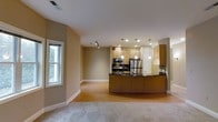 This single-floor space offers an open-concept layout that integrates the kitchen and living area into one cohesive and functional environment. The kitchen is a standout feature, showcasing light wood cabinetry, dark countertops, and a full suite of stainless steel appliances including a refrigerator, range, and built-in microwave. A curved kitchen island with a sink serves as both a practical workspace and a natural dividing point between the kitchen and the adjoining living area. Pendant lights hang above the island, adding warmth and focused illumination, while recessed lighting throughout ensures the space remains bright and welcoming. The living area transitions smoothly from hardwood-style flooring near the kitchen to plush carpeting, creating a comfortable and defined zone for relaxation. Neutral warm-toned walls contribute to a calm and inviting ambiance throughout. A window along the side wall allows natural light to filter in, enhancing the open feel of the space. Track lighting is also present, offering additional flexibility in how the space is lit. Overall, the layout is practical, modern, and well-suited for everyday living.