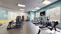 Located at 1767 Hermitage Blvd in Tallahassee, Florida, this fitness facility offers a comprehensive workout environment across a generously sized single-floor layout. The space is divided into two distinct areas: a fully equipped fitness room and an indoor racquetball court, connected via a glass-panel door that allows natural sightlines between the two zones.

The fitness room features a thoughtfully designed interior with light teal accent walls, bright recessed panel lighting, and large windows that invite ample natural light and outdoor views of the surrounding greenery. The durable rubber flooring supports a wide array of equipment, including treadmills, ellipticals, a stationary bike, strength training machines, and free weights. A wall-mounted flat-screen television and motivational wall decals — including "Mindset Is Everything" and "Stronger Every Day" — contribute to an energizing atmosphere. A water fountain is conveniently installed along one wall, and full-length mirrors enhance the sense of space throughout the strength training area.

The adjacent racquetball court features a polished hardwood floor with clearly marked red boundary lines, tall white walls, and a vibrant teal accent wall at the far end. Overhead panel lighting ensures consistent illumination across the court. A glass rear wall provides a transparent view into the fitness room, creating a connected and open feel throughout the entire facility.