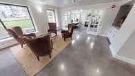Located at 319 N Tacoma Ave in Tacoma, Washington, this thoughtfully designed single-floor community space combines modern aesthetics with a warm, functional atmosphere. The entry lobby greets visitors with gleaming polished concrete floors, crisp white walls, and a built-in media console adorned with elegant chevron tile and marble-style cabinetry — setting a refined yet welcoming tone from the moment you step inside.

The lounge area is a standout feature, centered around a grouping of rich brown leather wingback chairs arranged around a woven jute rug and a round wooden coffee table. Large picture windows on multiple walls flood the space with natural light and offer views of the surrounding greenery and landscaped grounds, creating a bright and inviting atmosphere for gathering or relaxation.

French doors connect the lounge to a dedicated dining area, which continues the open, airy feel of the space. A private office is also available, finished with plush gray carpeting, a substantial dark wood desk, upholstered seating, and curated artwork — providing a professional and functional workspace.

Throughout, the design balances clean architectural lines with warm, residential touches, making this a versatile and polished space well-suited for community engagement and day-to-day operations.