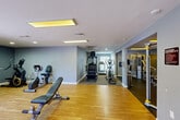 Located at 300 Woerner Road in Houston, TX, this single-floor fitness center offers a thoughtfully organized workout facility divided into two connected gym areas. The space features warm hardwood flooring in the main exercise zone, complemented by carpet-covered sections that provide a comfortable surface for cardio equipment use. Soft blue-gray walls create a calm, focused atmosphere throughout, while flush-mounted fluorescent light panels and recessed ceiling lights ensure bright, even illumination across the entire facility. The main gym area provides generous open floor space for free-weight training, featuring an adjustable weight bench and a multi-station Hoist cable machine. A full-length wall mirror enhances the sense of space and supports proper form during workouts. The secondary gym zone is well-stocked with cardio equipment, including a StairMaster, elliptical trainer, stationary bike, treadmill, and a rowing machine, alongside a medicine ball rack for functional training. A wall-mounted water fountain is conveniently located near the cardio area. Double French doors offer natural light at the entrance, and a dedicated storage closet provides space for additional equipment or supplies. The layout flows efficiently between zones, making this a versatile and fully equipped fitness facility.