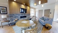 This single-floor lounge space combines contemporary design with warm, inviting materials to create a comfortable and functional gathering area. The room's most prominent feature is a sleek, linear fireplace framed by a floor-to-ceiling stacked stone surround, above which a large wall-mounted flat-screen television is set against rich, warm-toned wood cabinetry. This focal wall anchors the space and establishes its modern yet cozy aesthetic.

The ceiling transitions from a warm wood-paneled section above the fireplace wall to a higher, white-painted section over the seating area, adding architectural interest and a sense of openness. Pendant lighting fixtures hang at varying heights, complementing the recessed lighting to provide warm, layered illumination throughout.

A round dining table with lattice-backed chairs occupies the center of the room, offering a casual spot for dining or conversation. Adjacent to this, a generously sized gray sofa accented with blue and green throw pillows sits atop a patterned area rug, creating a relaxed lounge zone. Large windows along one wall flood the space with natural light and provide views to the exterior.

Hardwood-style flooring ties the entire space together, while the neutral color palette of grays, whites, and natural wood tones lends the room a clean, contemporary feel that is both functional and welcoming.