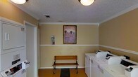 This single-floor space presents a functional yet thoughtfully decorated interior. The laundry area is equipped with a side-by-side washer and dryer, positioned along the right wall and finished in white. The room features warm beige walls accented by white crown molding and a chair-rail-style wainscoting detail that runs along the perimeter, adding a touch of classic architectural character.

A wooden bench with a wrought-iron frame is centrally placed against the back wall, offering a practical seating option. Above it hangs a framed painting depicting a scenic landscape with hot air balloons, providing a decorative focal point. A fire extinguisher mounted on the left wall reflects attention to safety. The floor is covered in a warm-toned surface, and a dark entry mat is positioned in front of the bench.

Overhead, a flush-mount ceiling light fixture provides ambient illumination, complemented by a textured ceiling. A ventilation grille is also visible, indicating climate control provisions. Overall, the space balances utility with simple decorative touches, creating a tidy and organized environment.