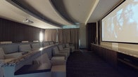 This refined screening room is designed with a strong emphasis on both functionality and aesthetic sophistication. The space is anchored by a large projection screen positioned along one wall, framed by rich dark wood paneling that extends throughout the room and contributes to its intimate, immersive atmosphere. A distinctive architectural feature is the curved, tiered ceiling with recessed cove lighting that casts a soft ambient glow, reinforcing the room's elegant character.

Seating is arranged in a gently curved configuration, consisting of generously proportioned cream-colored upholstered armchairs, each paired with sleek side tables featuring marble-like surfaces. This layout ensures unobstructed sightlines to the screen from every seat. The chairs are arranged in a sweeping arc that follows the room's curved design language, creating a cohesive and carefully considered layout.

Floor-to-ceiling drapes in warm champagne tones line one side of the room, adding texture while contributing to the acoustic and visual warmth of the space. A granite-topped credenza runs along the screen wall, providing functional surface space. Dark, patterned carpeting grounds the room and complements the overall color palette of neutral tones, dark wood, and soft lighting, resulting in a polished, theater-quality environment.
