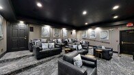 Located in Charlotte, NC, this elegantly designed home theater room delivers an immersive cinematic experience within a well-appointed, single-level space. The room is anchored by a large wall-mounted flat-screen display, framed within a dark, custom-built recessed panel that commands immediate visual attention upon entry.

The interior follows a refined dark palette, with deep charcoal ceilings and warm gray walls creating an intimate, theater-like ambiance. Recessed lighting is strategically placed throughout to provide subtle, even illumination without disrupting the cinematic atmosphere. The flooring features a bold gray marble-patterned surface that adds texture and visual depth to the room.

Seating is arranged in a tiered configuration, accommodating multiple viewing rows for an authentic theater experience. Dark charcoal leather sofas and loveseats are dressed with blue and gray accent pillows, offering both comfort and style. Compact gold-toned side tables are positioned throughout for added functionality and a touch of warmth against the cool tones of the room.

The rear wall serves as a gallery-style backdrop, adorned with a series of four framed abstract artworks in blue and white hues, illuminated by wall-mounted accent lighting. A sleek black handrail guides movement between the tiered levels. The overall design is cohesive, sophisticated, and purpose-built for premium entertainment.